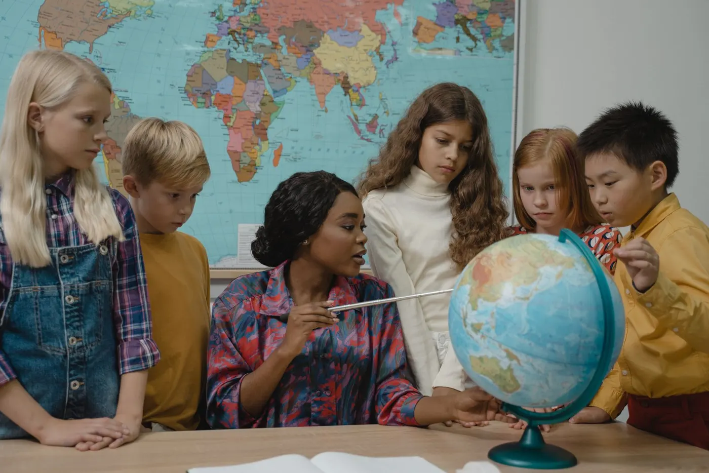 A teacher and children from many backgrounds exploring a globe in front of a world map